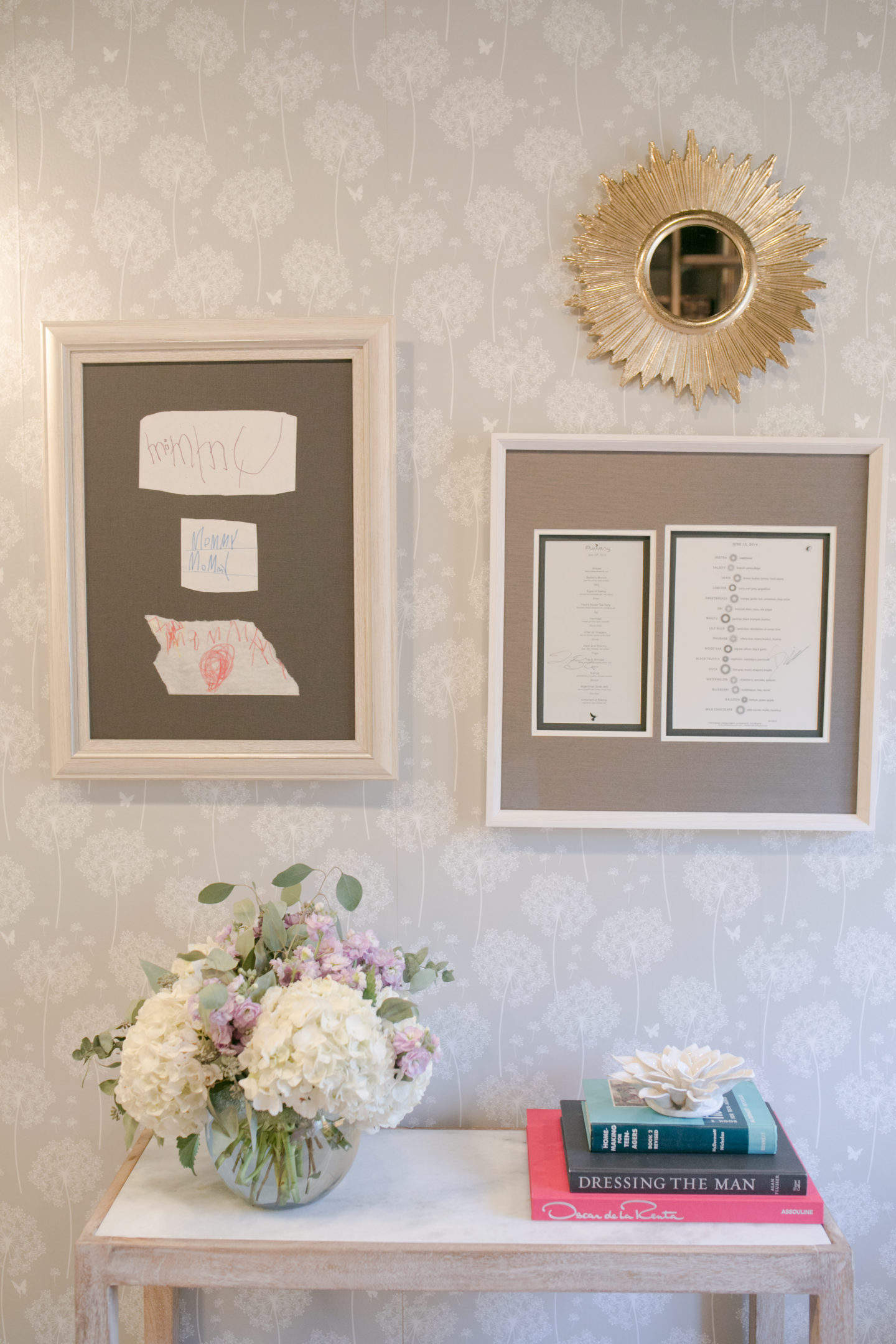 Wall pops wallpaper dandelion wallpaper hung with personal art and a gold starburst mirror and hydrangea floral arrangement on a white console table