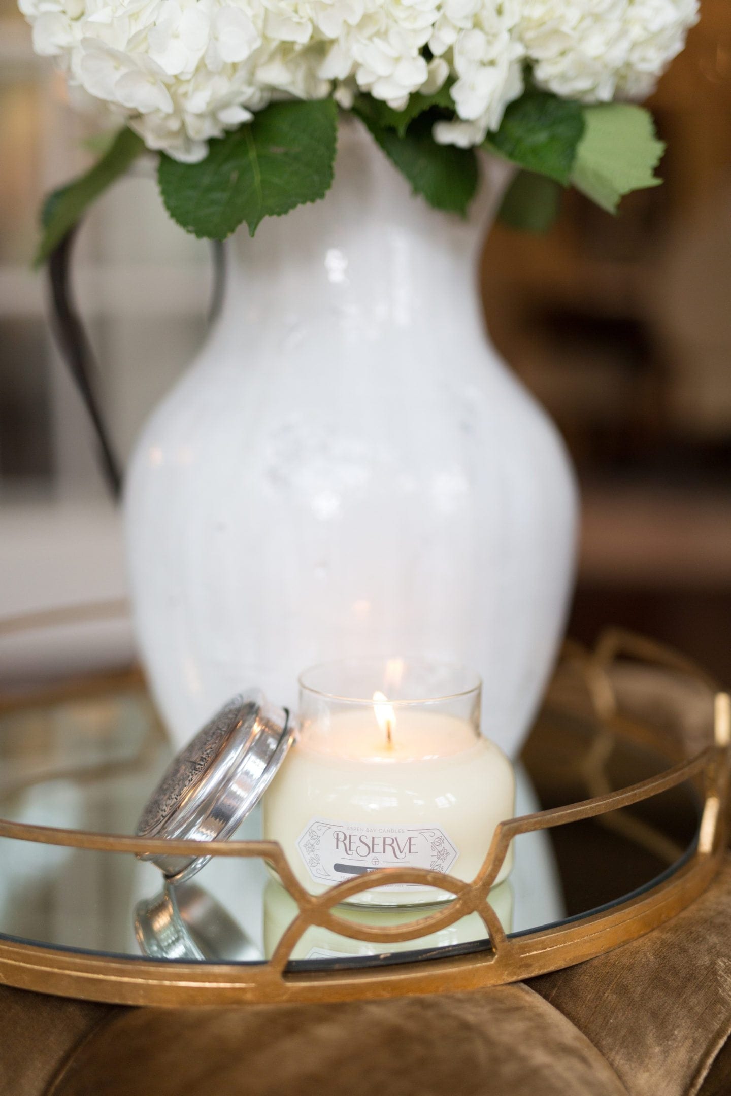 How to decorate a tray in a living room. I use our gold metallic round tray to display flowers and leave a candle to warm up a living room. Just these two easy tricks make a room feel comfortable. The Tuscan Urn is filled with hydrangeas and looks beautiful on the velvet ottoman and tray even without the flowers!
