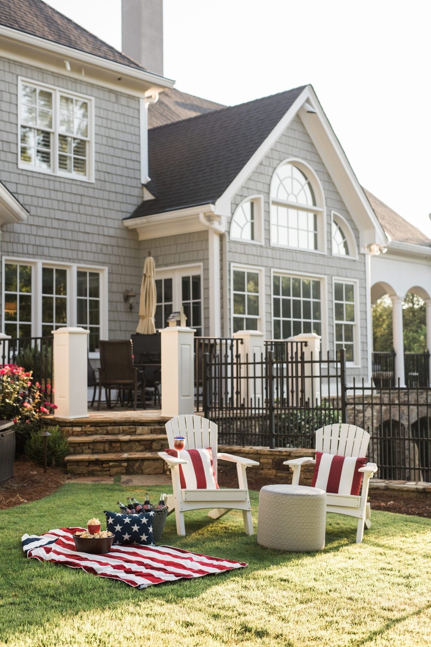White wood adirondack chairs with red and white strip outdoor pillows set the stage for a Fourth of July backyard party. Gray shingle house Cape Cod style house with Americana felt an outdoor porch setting.