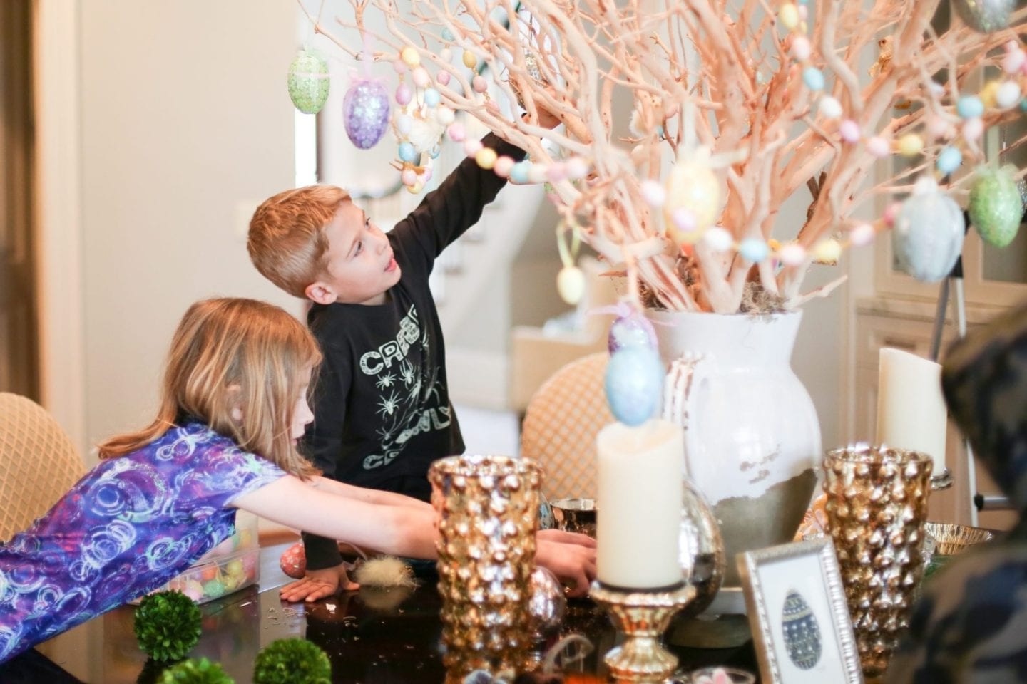 Kids decorating for Easter with Easter egg garland and Egg ornaments handing from branch centerpiece. 