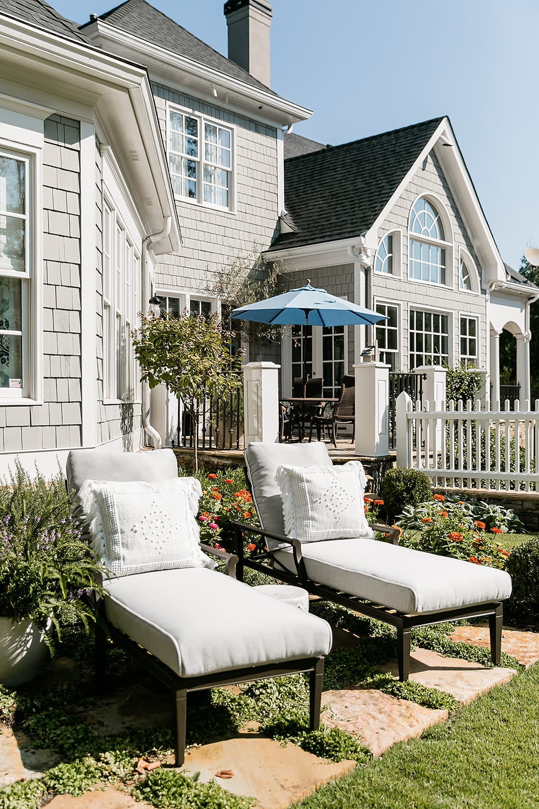 Woodard Furniture. Chaise lounges in Andover Collection. Outdoor furniture in light gray. White and gray cedar home exterior with white picket fence and blue outdoor umbrella.