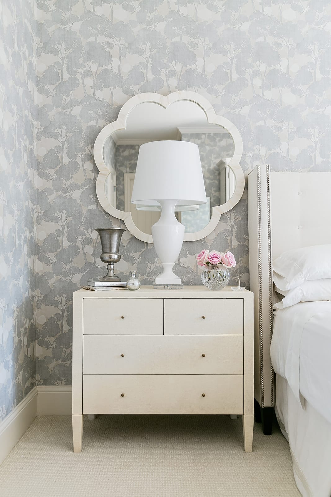 Bedroom Nightstand Decor with ivory quatrefoil mirror, white desk lamp and soft pink flowers with light lavender floral wallpaper.