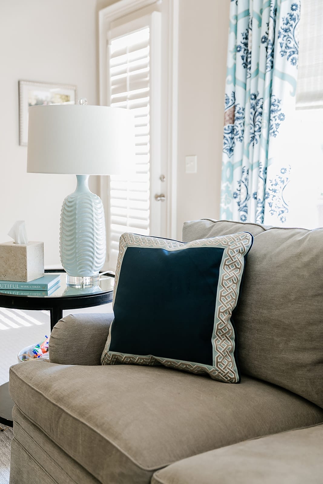 Light blue lamp in family room. Blue drapes, navy blue velvet pillow and taupe couch.