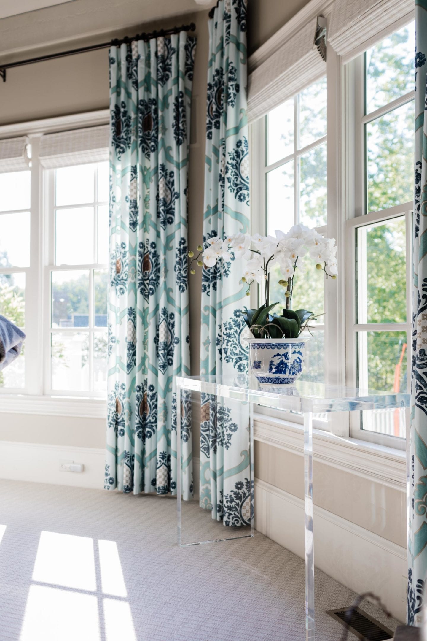 Lifestyle blogger Kelly Page for BlueGrayGal shares how she styles a fake orchid on an acrylic console table. This Frontgate orchid has blue and white ceramic pot with white flowers surrounded by blue draperies. The faux orchid plant sits in a living room setting for easy plant care.