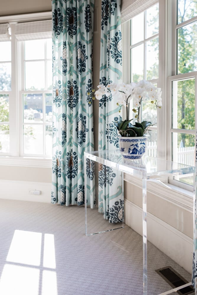Lifestyle blogger Kelly Page for BlueGrayGal shares how she styles a fake orchid on an acrylic console table. This Frontgate orchid has blue and white ceramic pot with white flowers surrounded by blue draperies. The faux orchid plant sits in a living room setting for easy plant care.