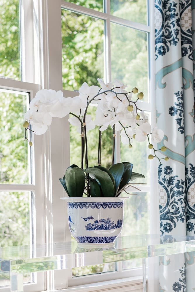 Lifestyle blogger Kelly Page for BlueGrayGal shares how she styles a fake orchid on an acrylic console table. This Frontgate orchid has blue and white ceramic pot with white flowers surrounded by blue draperies. This Frontgate faux orchid plant sits in a living room setting for easy plant care.