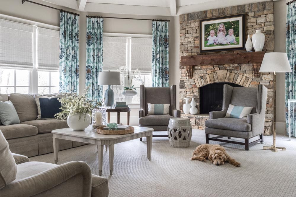 Traditional family room with stacked stone fireplace is framed with a tweed sitting area, ivory coffee table and a neutral soft layered with custom designer throw pillows.