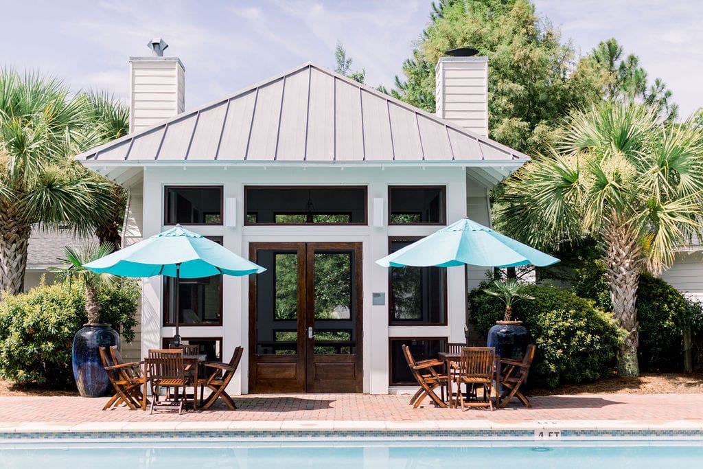A summer kitchen makes for this pool house in white with dark stained doors. The teak furniture has bright blue outdoor umbrellas.