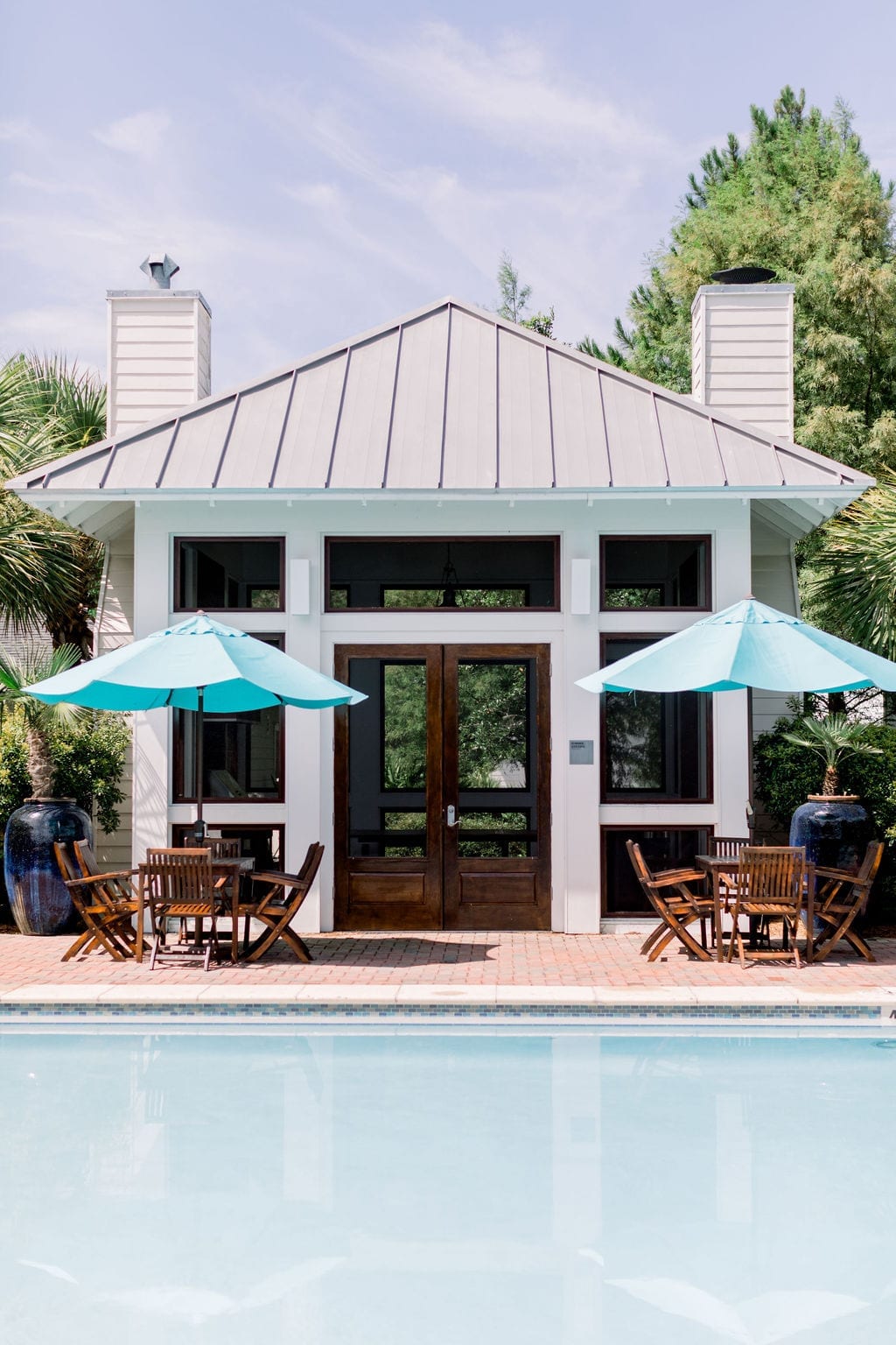 A summer kitchen makes for this pool house in white with dark stained doors. The teak furniture has bright blue outdoor umbrellas.