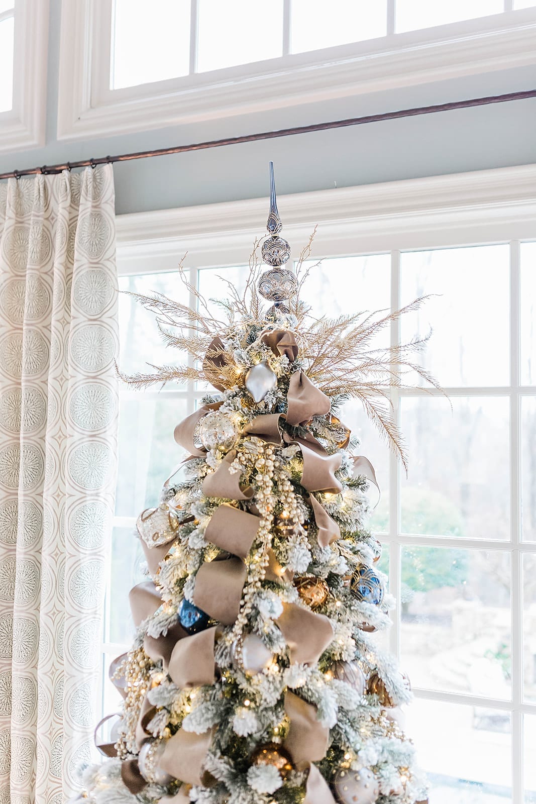 A Taupe velvet ribbon Christmas tree with blue ornaments from Frontgate collection and Frontgate tree topper. A gold Christmas skirt finishes off the taupe and navy blue accented flocked Christmas tree.