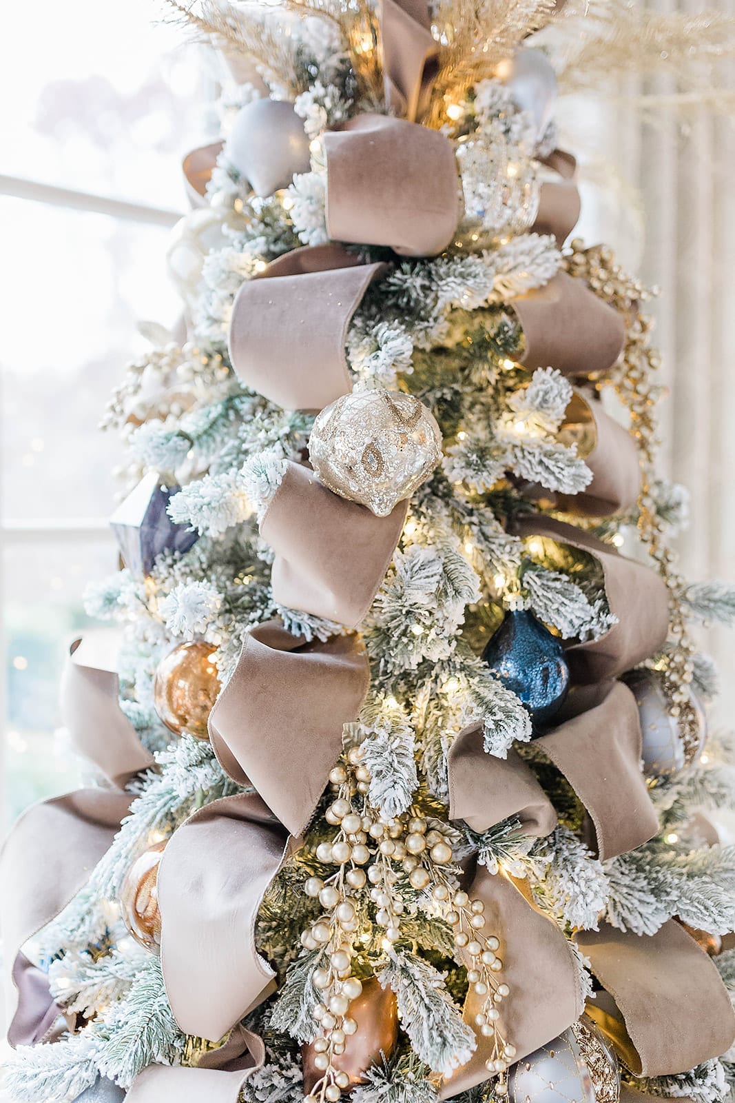 A Taupe velvet ribbon Christmas tree with blue ornaments from Frontgate collection and Frontgate tree topper. A gold Christmas skirt finishes off the taupe and navy blue accented flocked Christmas tree.