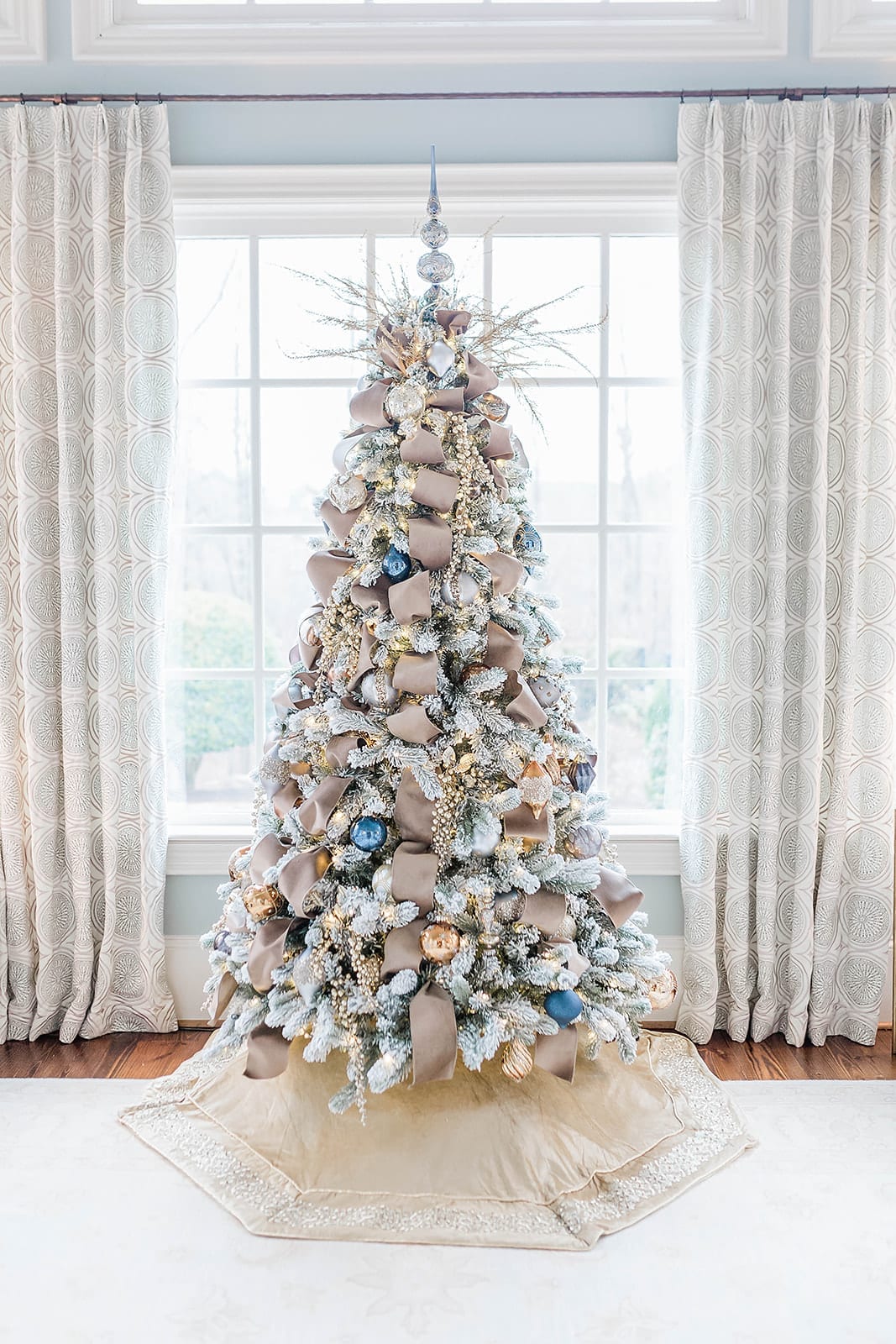 A Taupe velvet ribbon Christmas tree with blue ornaments from Frontgate collection and Frontgate tree topper. A gold Christmas skirt finishes off the taupe and navy blue accented flocked Christmas tree.