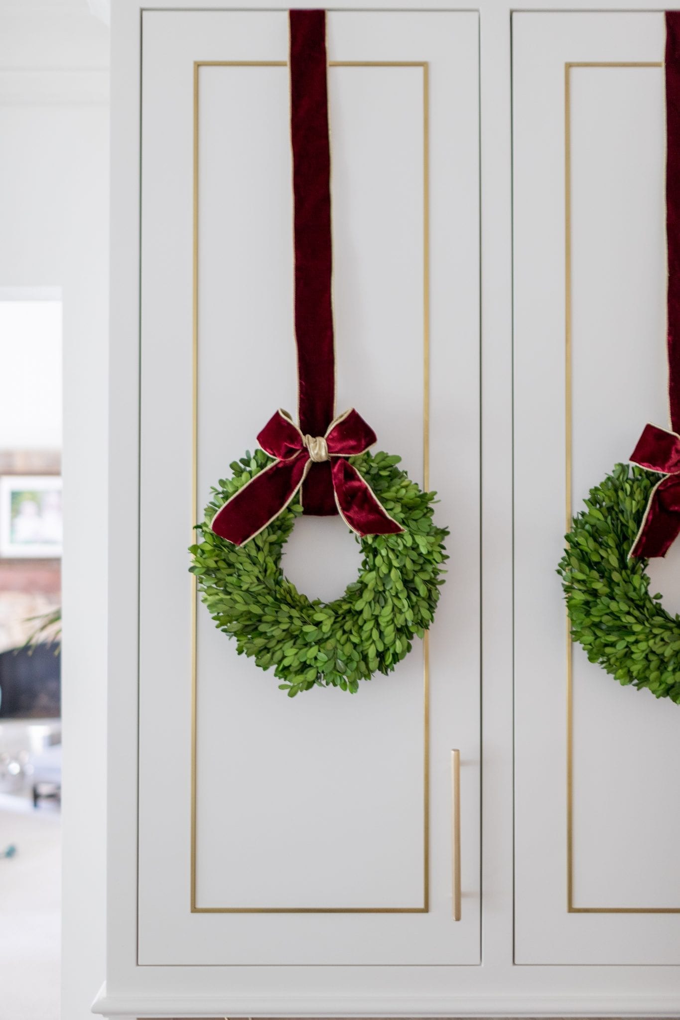 Preserved boxwood wreath hung with burgundy velvet ribbon on a white and gold kitchen cabinet.