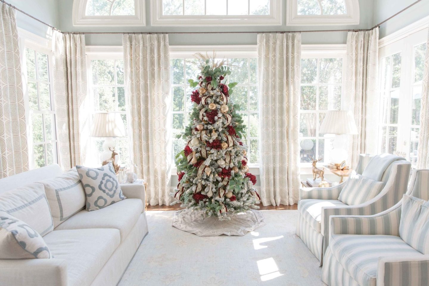 Red and gold flocked Christmas tree in a light blue sunroom with green tree sprays.
